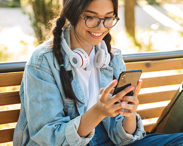 positive young student wearing eyeglasses sitting on bench outdoors in nature park using mobile phone.